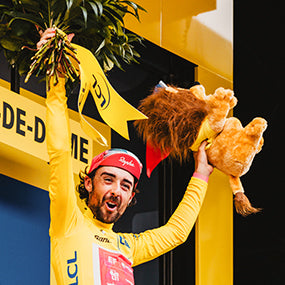 A rider in the yellow leader's jersey celebrates on the podium with a bouquet and a lion mascot