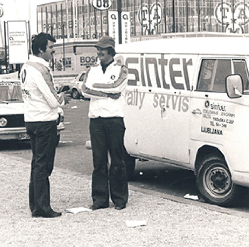 Black-and-white photo of two men talking beside a Sinter rally service van